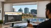 Business owner researching a custom blinds company in Barrie, Ontario, on a computer, with a waterfront city view and window blinds visible through large office windows